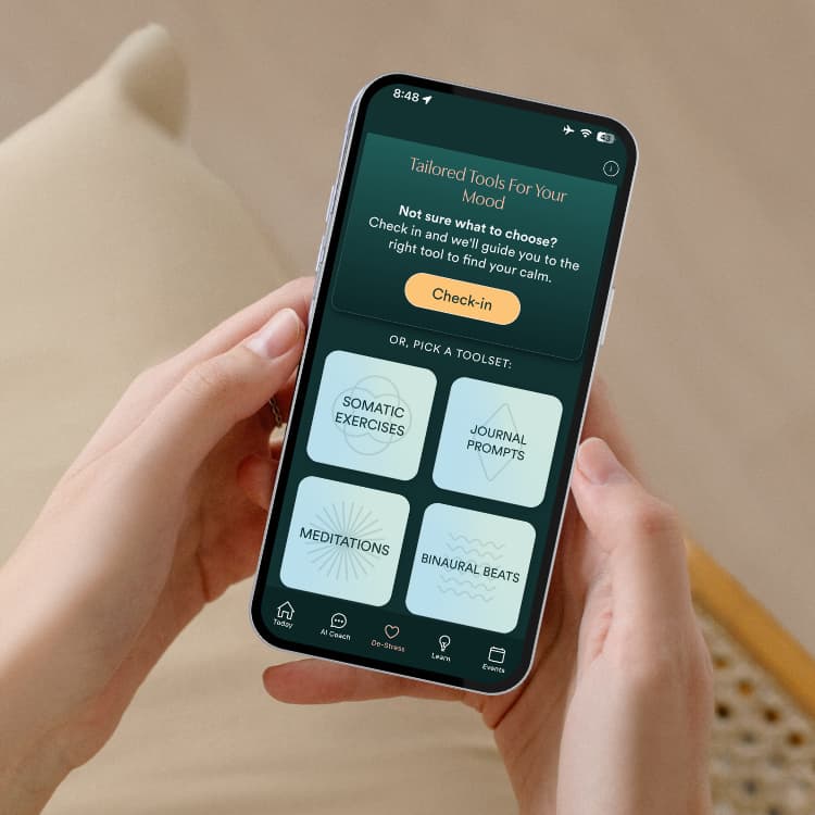 Person holds a smartphone showing a mood tool app with a prominent Check-in button and four tiles: Somatic Exercises, Journal Prompts, Meditations, and Binaural Beats.