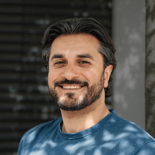Smiling man with dark hair and a beard wearing a blue shirt, standing outdoors near a stone wall.
