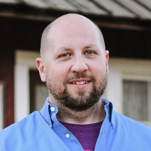 Head-and-shoulders portrait of a smiling bald man with a beard, wearing a blue shirt, standing outdoors in front of a house.