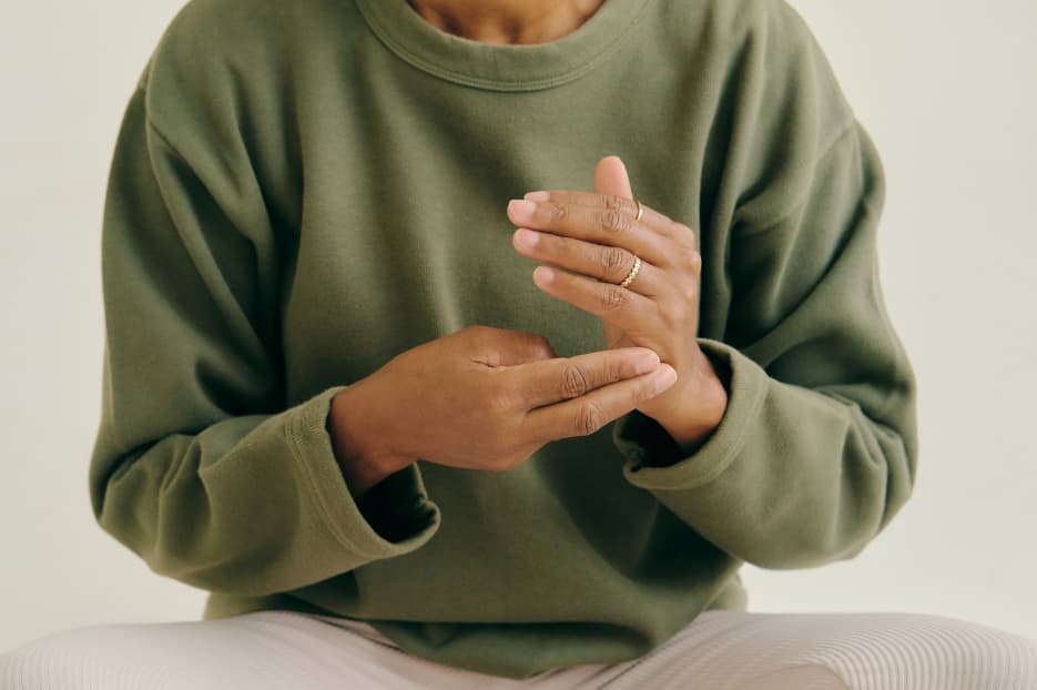 Person in an olive-green sweatshirt has palms pressed together in a prayer-like gesture against a light background.