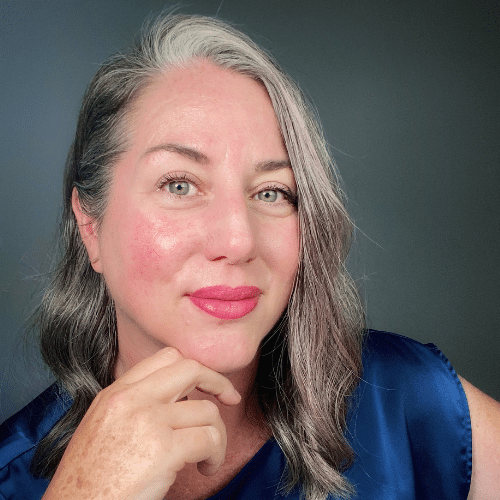 Close-up portrait of a smiling woman with gray hair, pink lipstick, and a blue blouse, resting her chin on her hand.