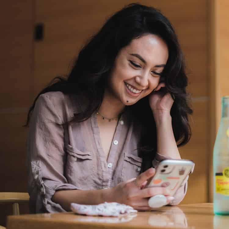 Woman with long dark hair smiling while looking at her smartphone at a wooden table, wearing a light pink blouse.