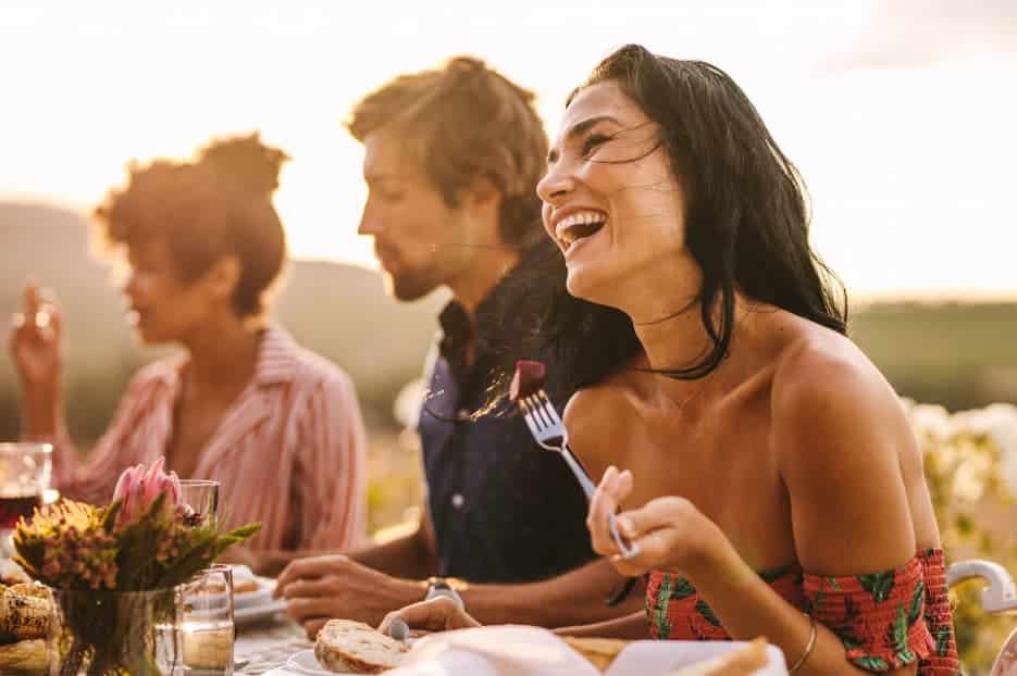 Group of friends enjoying an outdoor dinner at sunset; woman in the foreground laughs with a fork in hand.