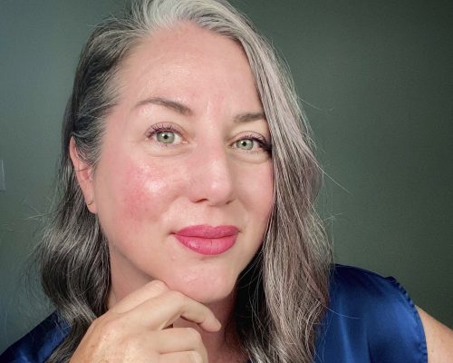 A woman with shoulder-length gray hair and pink lips poses for a close-up portrait, hand resting under her chin in a blue top.